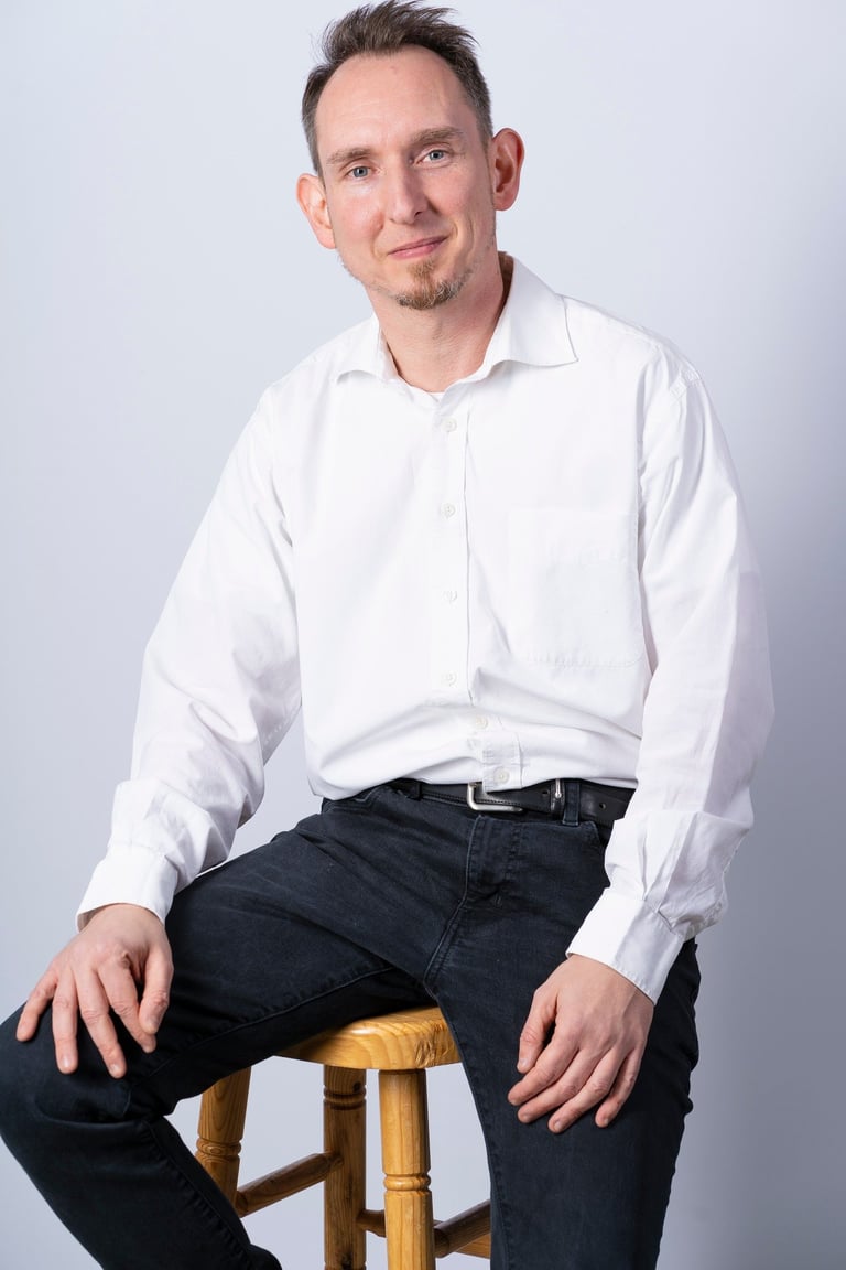 Man in white dress shirt and dark jeans sitting on wooden stool against light background, professional headshot style portrait